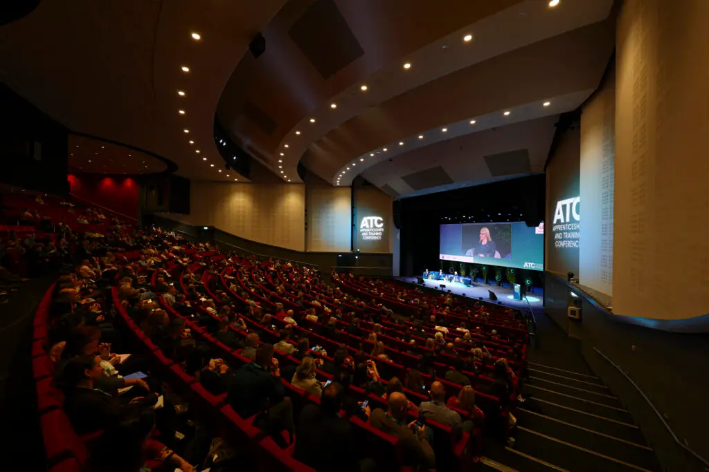 Large lecture hall filled with people watching a speaker on stage at an ATC conference work session or plenary. The stage is illuminated with screens and lighting, and the audience sits in red seats.5B5 Main stage at the front with presenters and a large display screen showing ATC branding.
