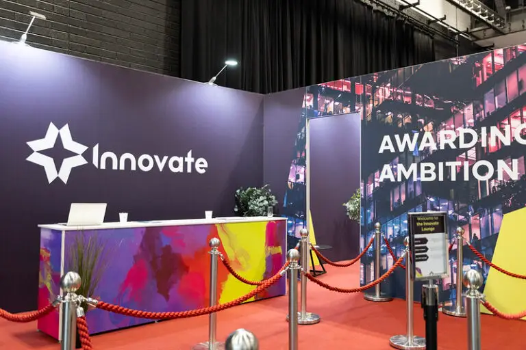 Trade show booth with a purple wall featuring the 'Innovate' logo, a colorful front desk, and red rope barriers under an 'Awarding Ambition' backdrop.