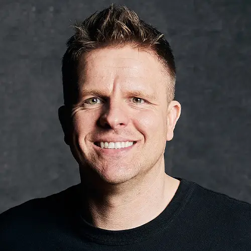 Portrait of a smiling man with short brown hair wearing a black shirt against a dark background.