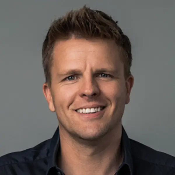 Head-and-shoulders portrait of a smiling man with short brown hair wearing a dark shirt against a neutral gray background