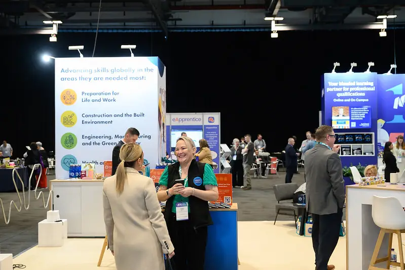 Two women converse at a trade show booth with a large informational display behind them and other attendees in the background.