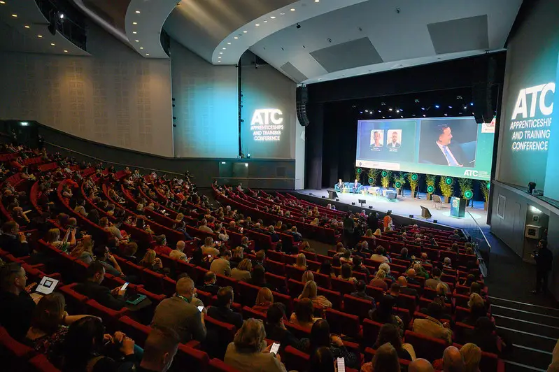 Large auditorium at an apprenticeship and training conference, with a stage, panelists on screen, and rows of attendees in red seats.