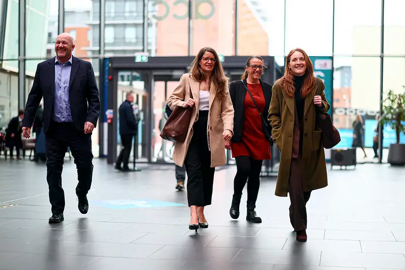 Four people walking together in a modern glass atrium, smiling and wearing business-casual coats and bags, mid-day light streaming in.