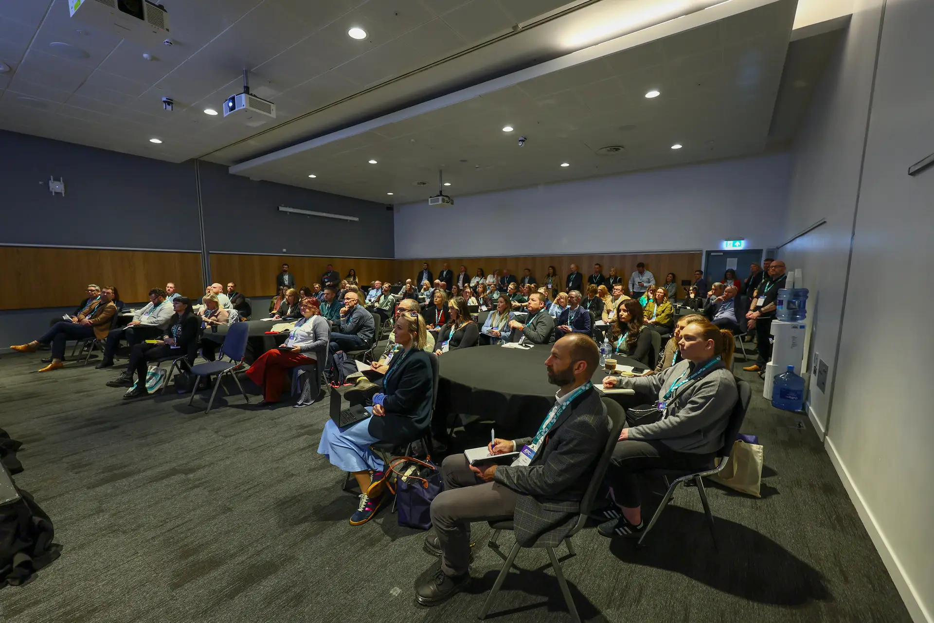 Audience of professionals seated in a conference room, facing the stage during a presentation.