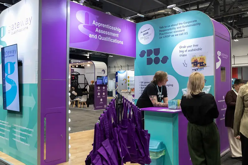 Trade show booth for apprenticeship assessment and qualifications, purple and turquoise display with a counter and attendees talking nearby.