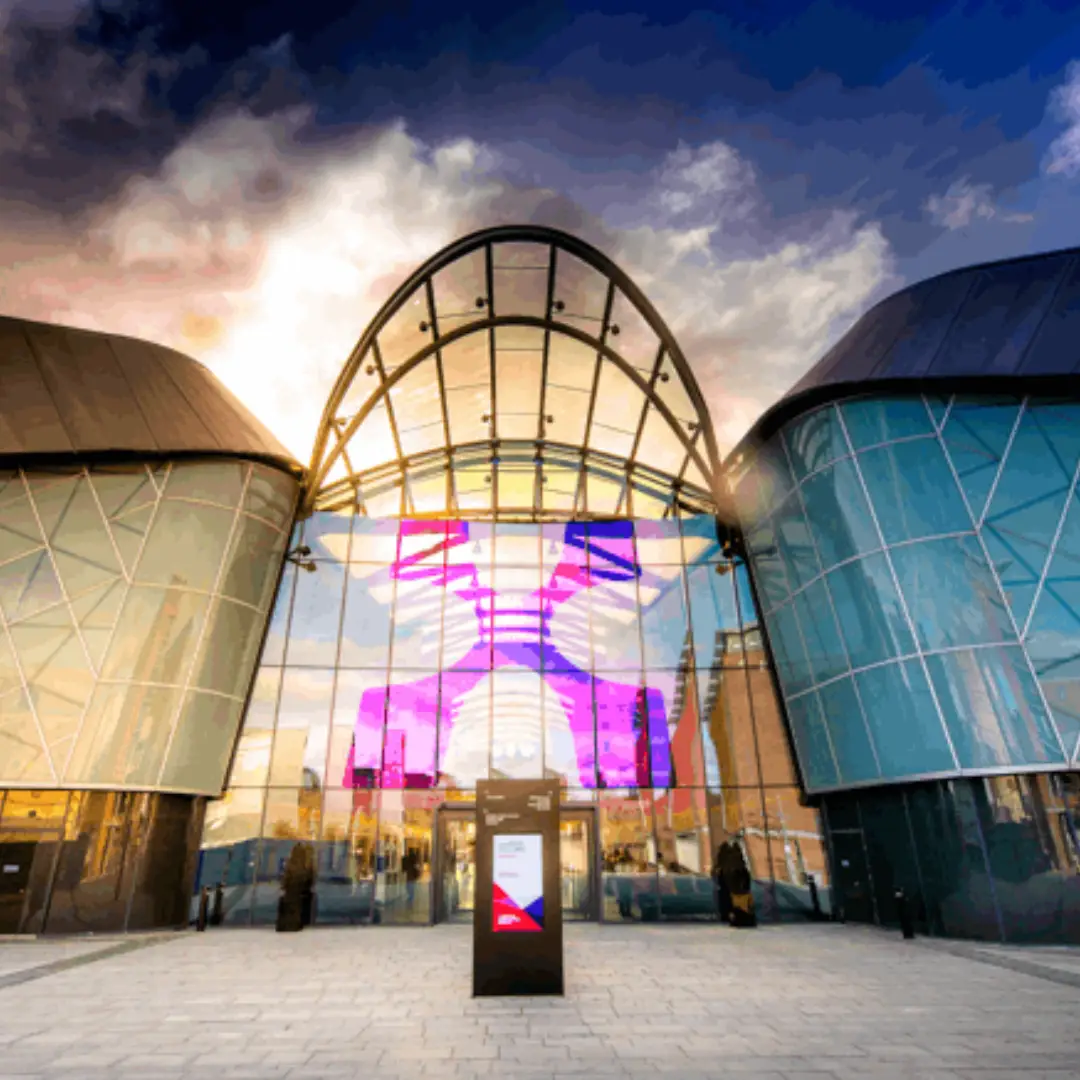 Contemporary glass building with a tall arched entrance and colorful digital display reflected in the facade, outdoor plaza in front.