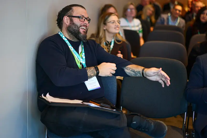 Man with a beard and glasses sits on a chair by a wall, gesturing with his right arm during a conference with other attendees in the background.