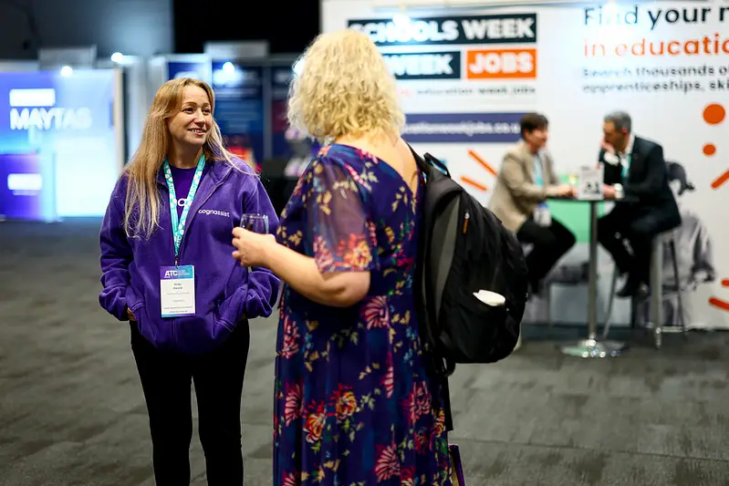 Woman in a purple jacket with a conference badge chats with a blonde visitor in a floral dress at a busy expo booth in the background.