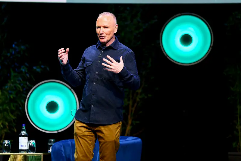 Male speaker on stage delivering a talk, hands gesturing, with teal circular lights behind him or in the background.
