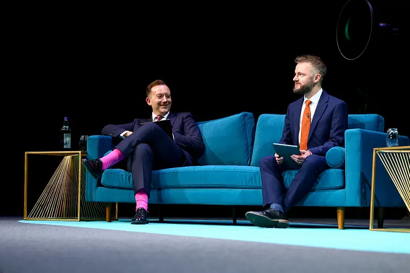 Two men in suits sit on a teal sofa on a stage, one with a tablet in lap and the other holding a notebook, mid-conversation on stage.
